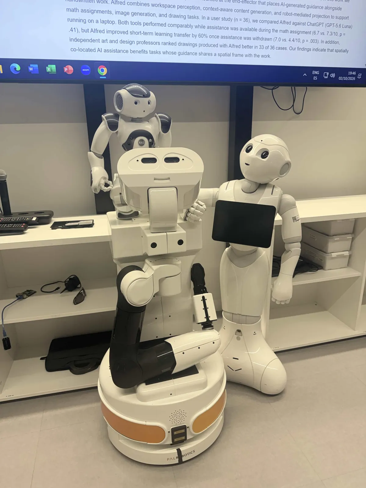 Robots in the IE Robotics and AI Lab, including a Pepper and a NAO humanoid, with the AIfred paper on screen behind.