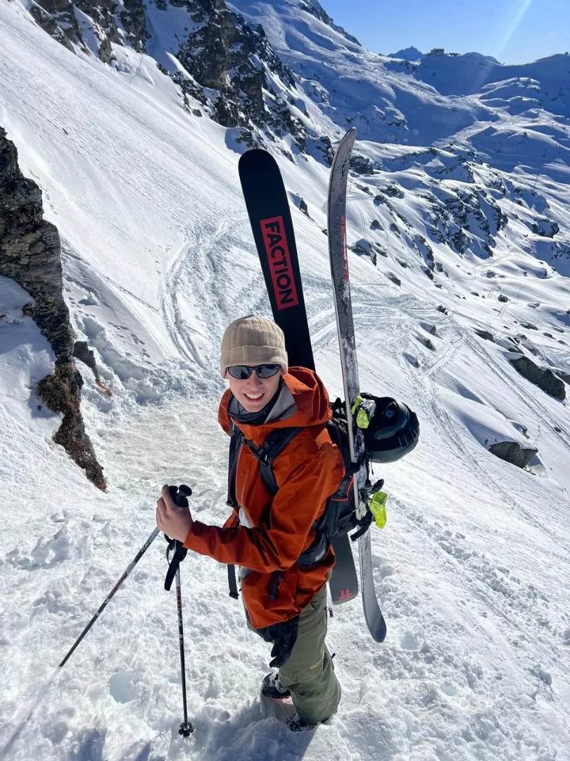 Gregorio Orlando in an orange jacket carrying skis up a steep snow slope.