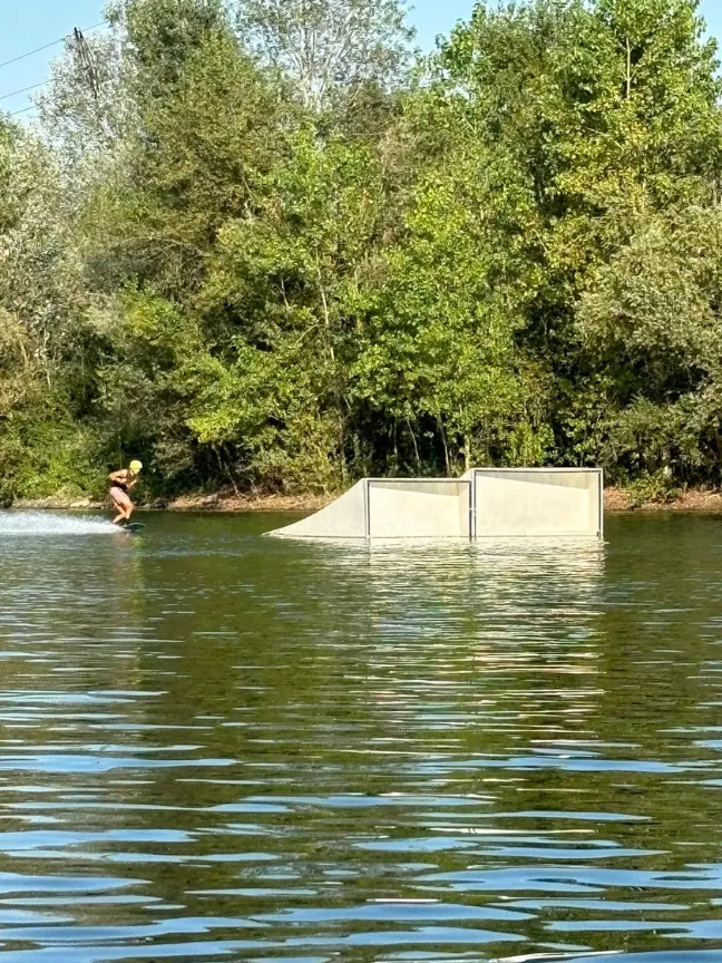 A wakeboarder riding towards a ramp on a lake surrounded by trees.